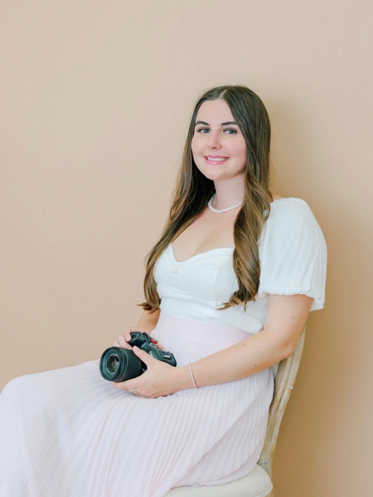 Pregnant photographer in white dress sitting in chair, smiling and holding a DSLR camera.
