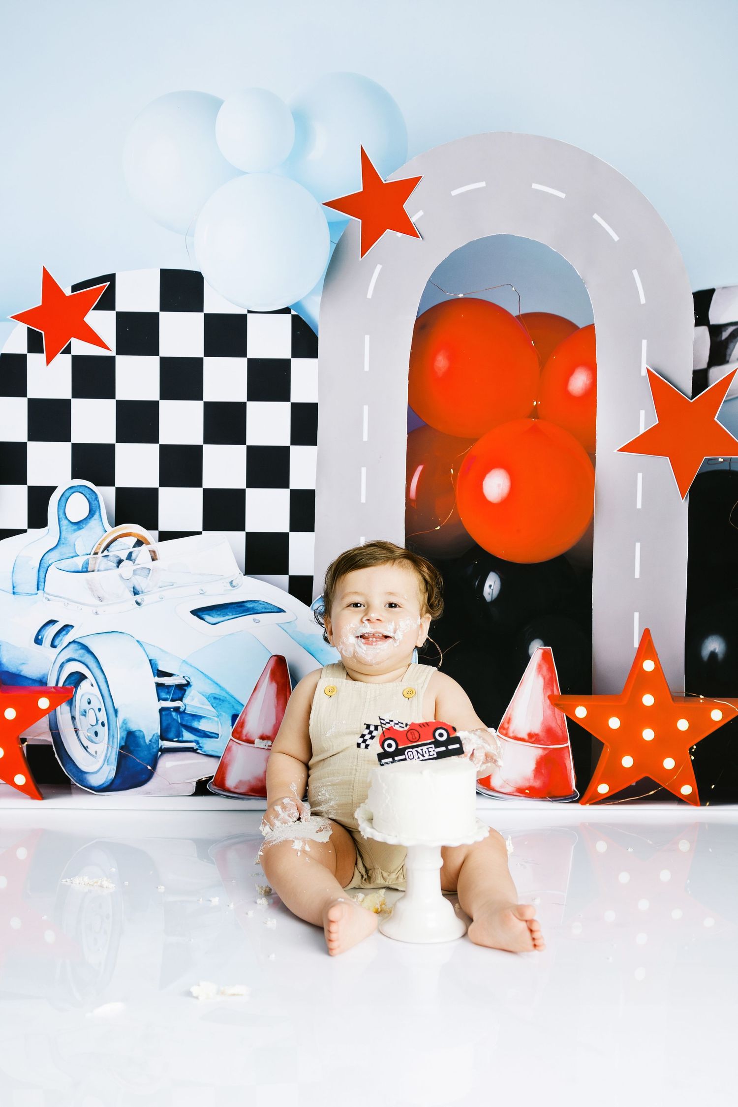 Baby smiles at cake smash birthday party with race car theme, balloons, and checkered flag backdrop.
