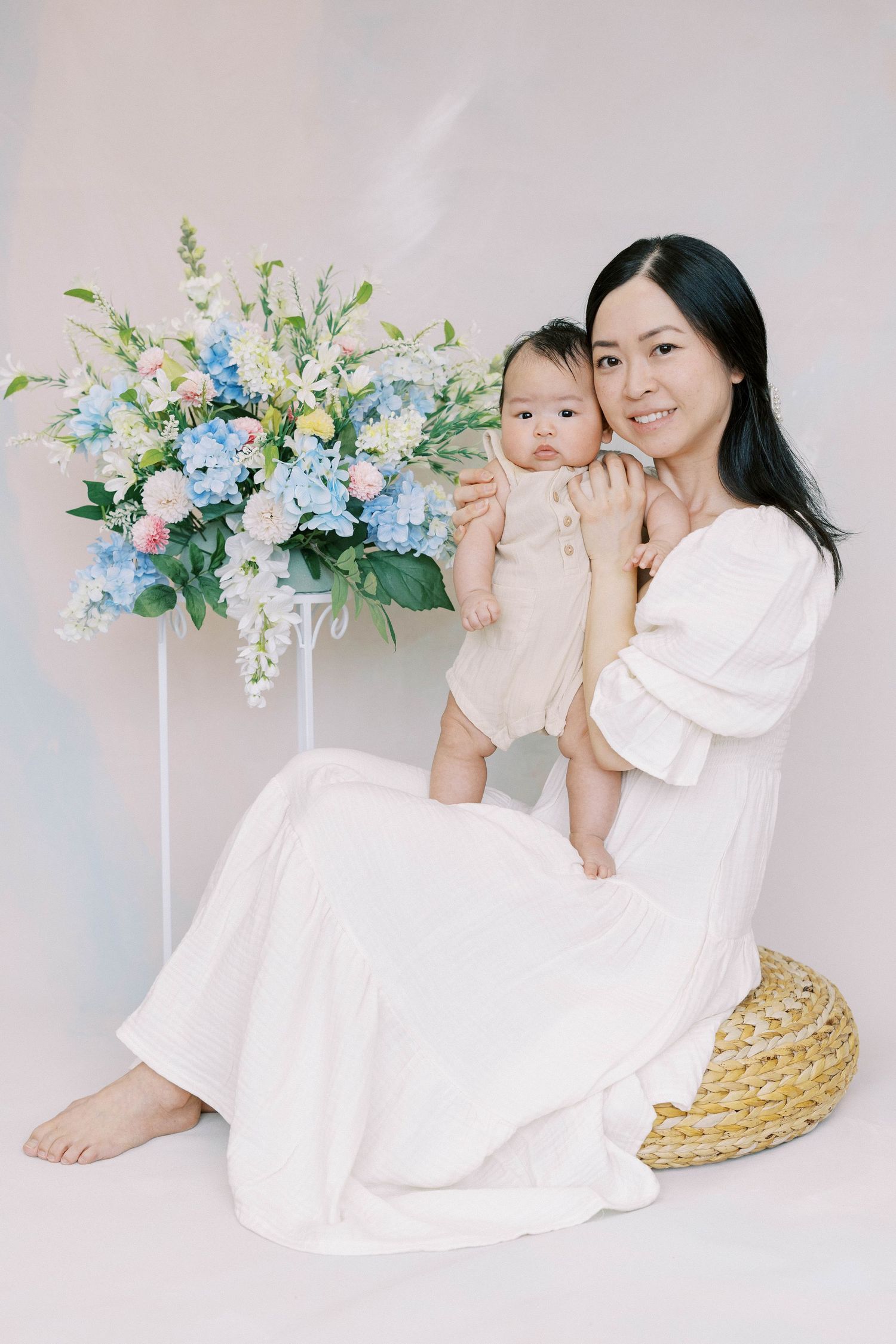 Asian mother in white dress smiling with baby on lap beside elegant blue and pink flower arrangement in studio.