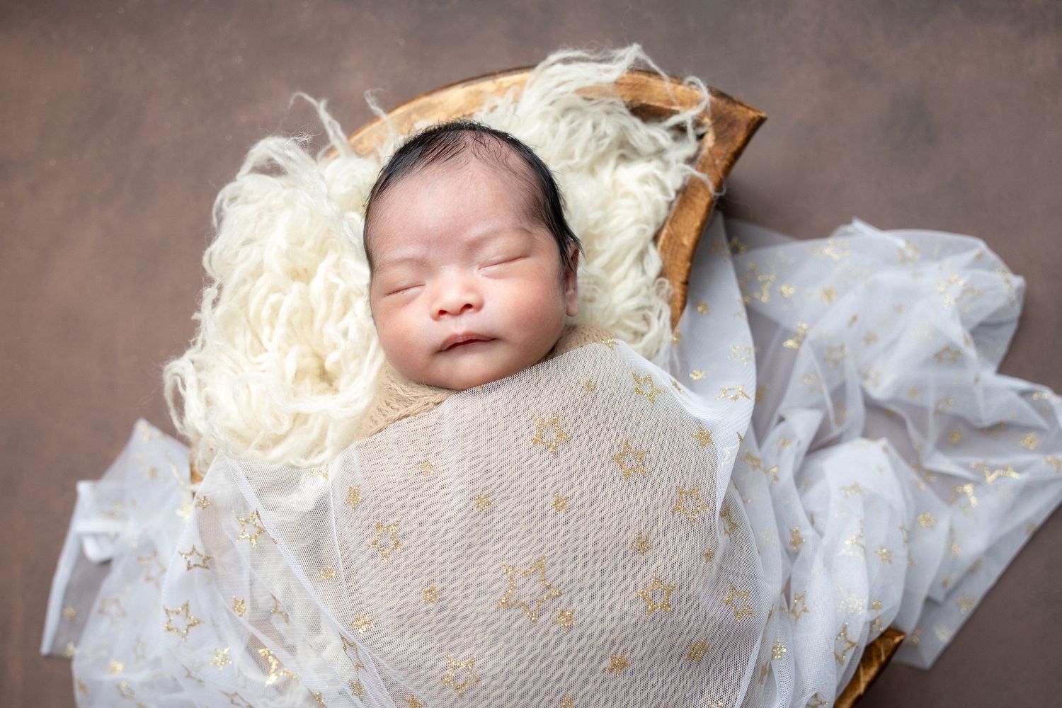 Sleeping newborn swaddled in cream wrap with gold details in wooden bowl on brown background.