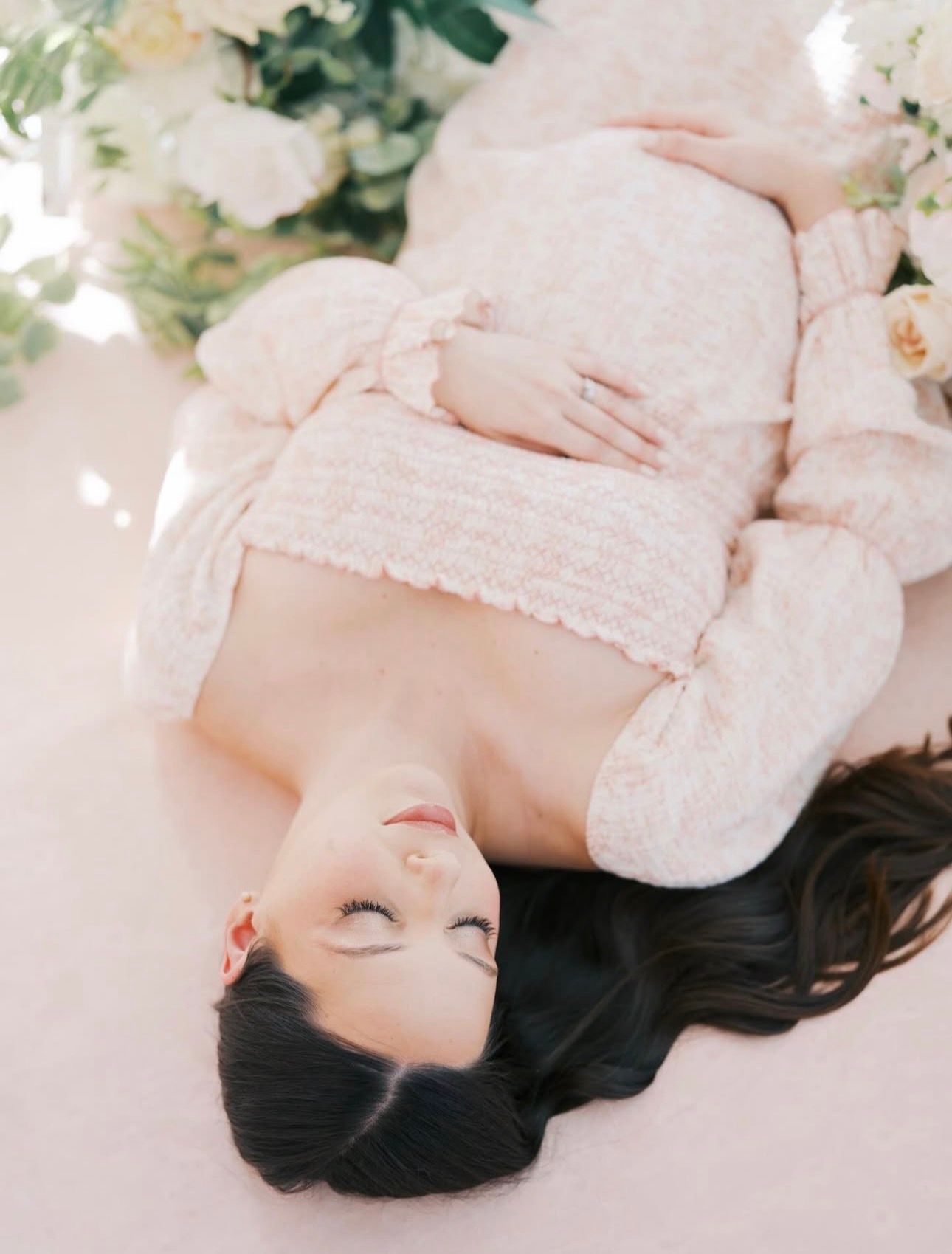 Pregnant woman in pink tweed outfit lying among white flowers with eyes closed in serene maternity photo.