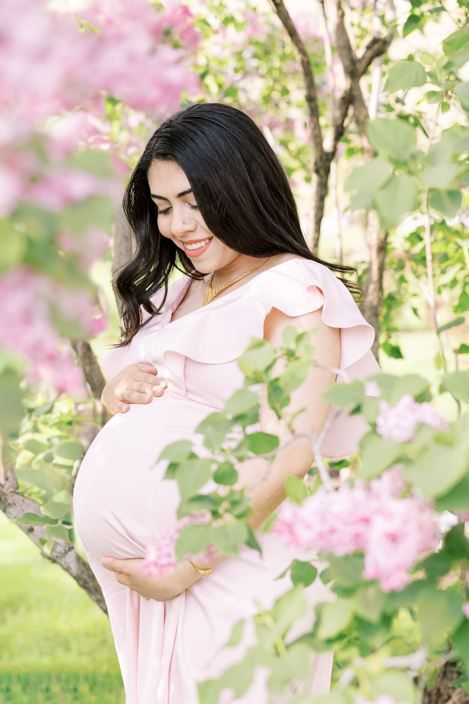 Glowing pregnant woman in blush dress gently holding baby bump surrounded by pink blooming flowers.