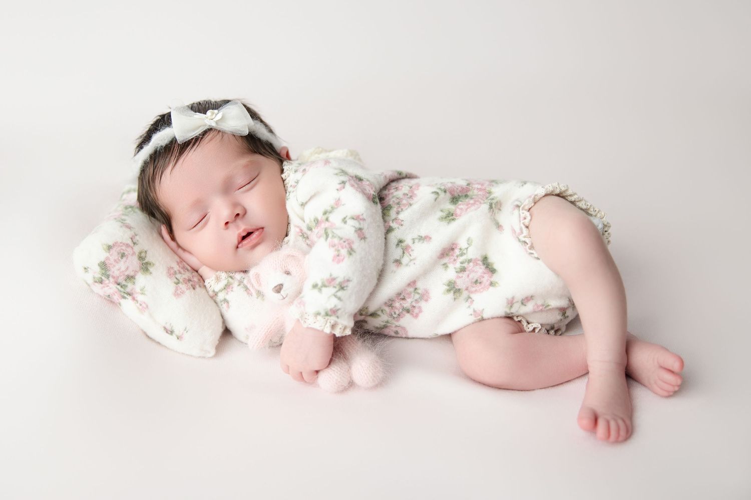 A peaceful newborn baby girl in a floral romper sleeps on a white pillow holding a tiny stuffed bunny.