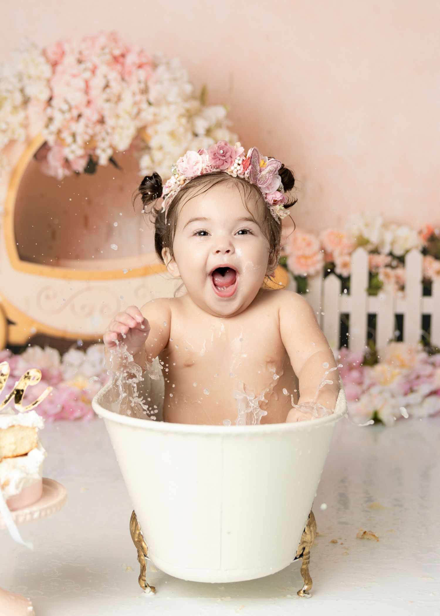 Laughing baby girl in floral crown splashing in white mini bathtub during first birthday cake smash photo session.