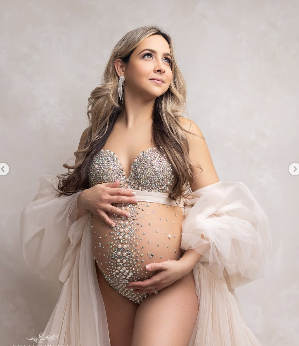 Pregnant woman in jeweled maternity bodysuit with sheer robe, posing elegantly in professional studio photo.