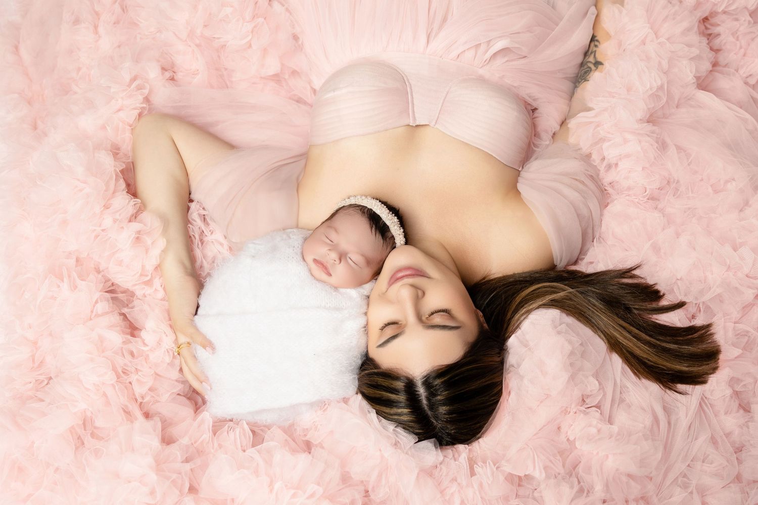 A mother lies peacefully with her newborn baby on a soft pink fluffy backdrop, both dressed in delicate pink.