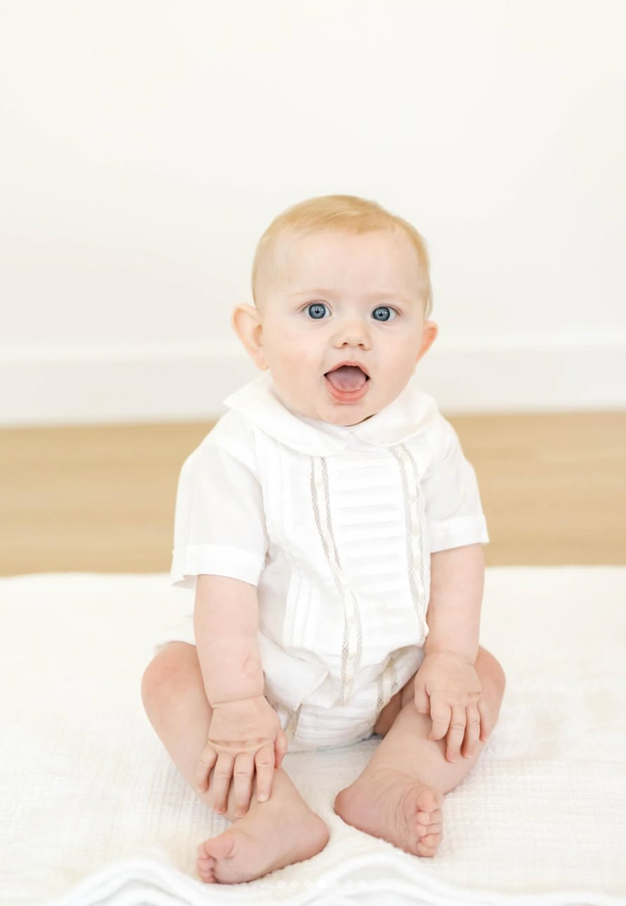 Adorable blue-eyed baby in white outfit sitting upright with mouth open on a white blanket.