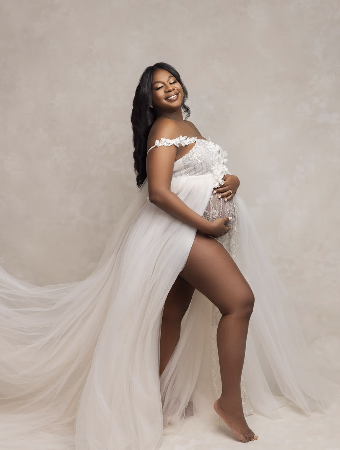 Pregnant woman in elegant white off-shoulder tulle gown cradling baby bump in a professional maternity studio photoshoot.