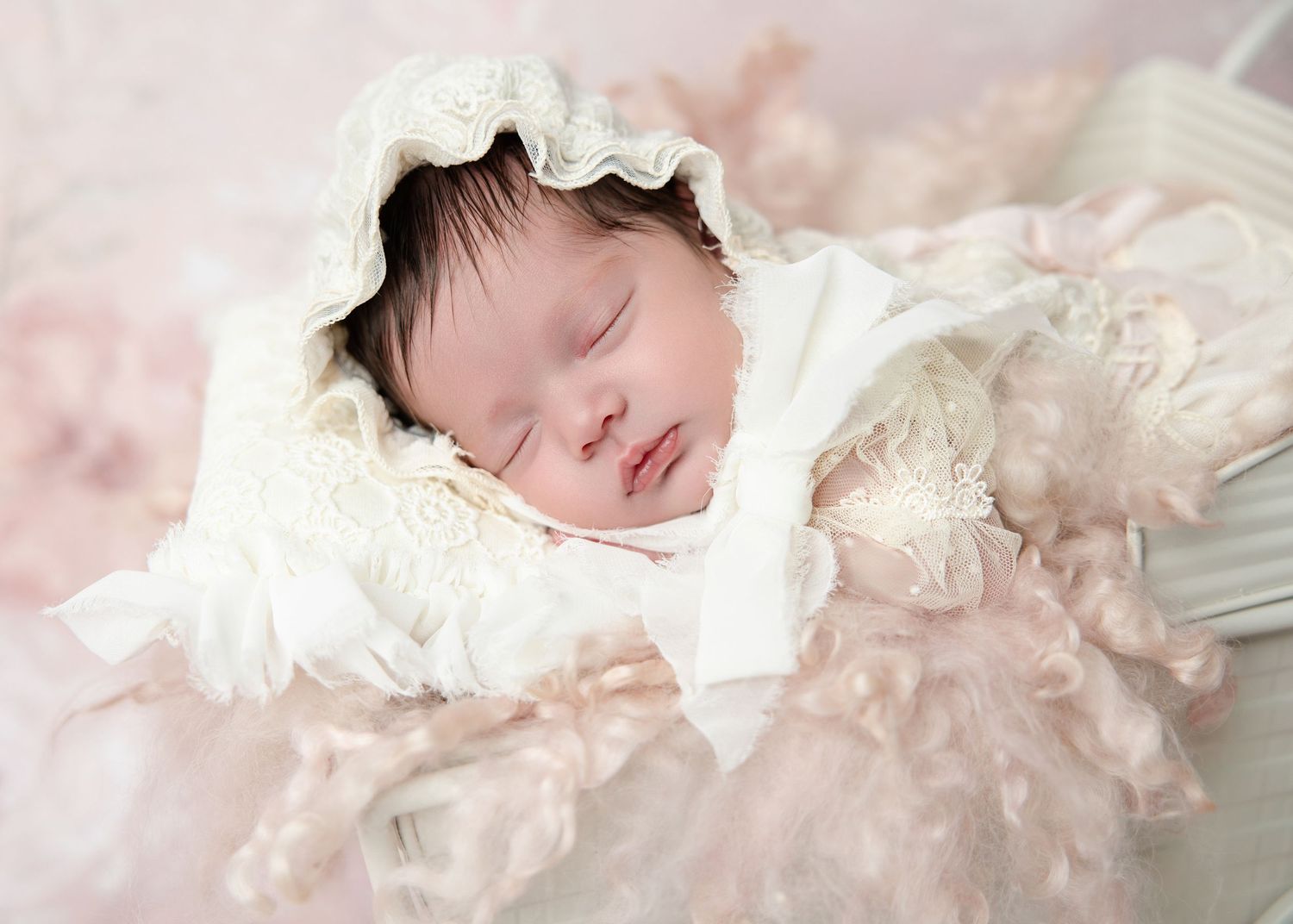A close-up of a sleeping newborn baby wearing a delicate lace bonnet surrounded by soft pink ruffles.