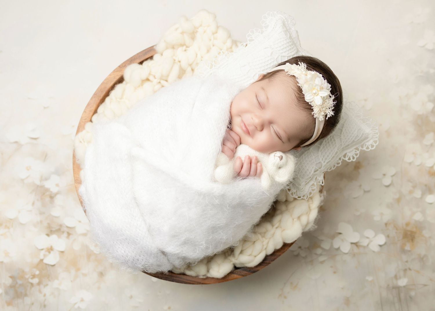 Sleeping newborn swaddled in white wrap with floral headband in wooden bowl prop.