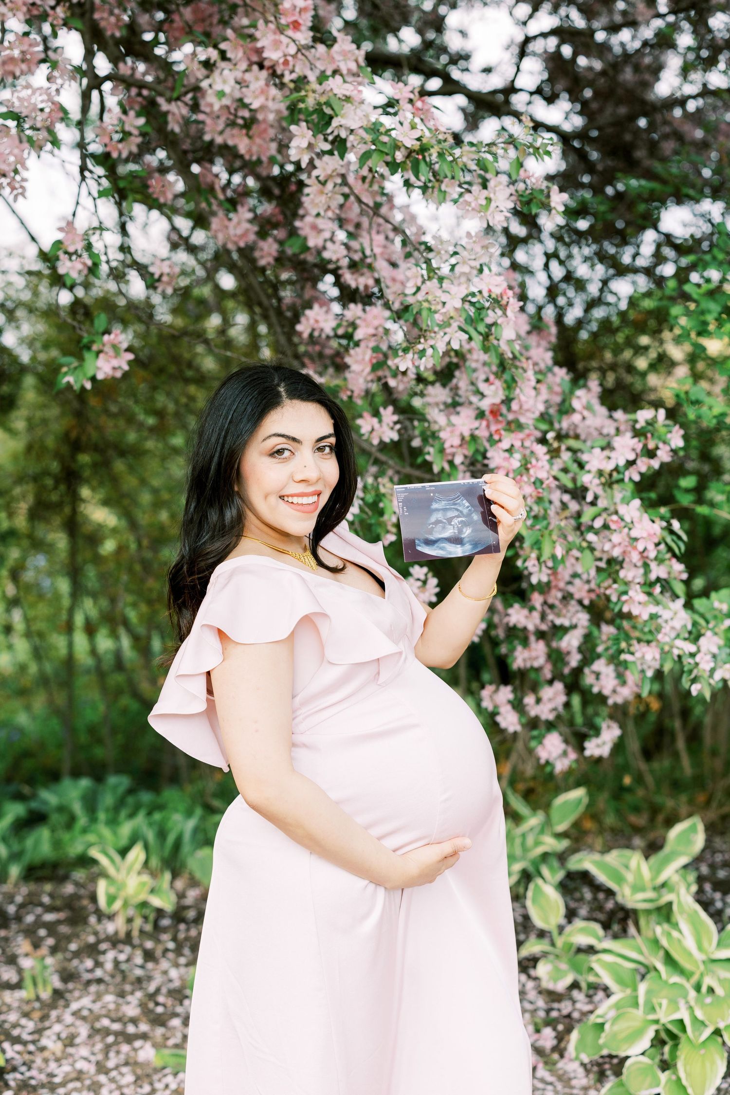 Smiling pregnant woman in pink dress holds ultrasound photo beside blooming pink flowering tree.