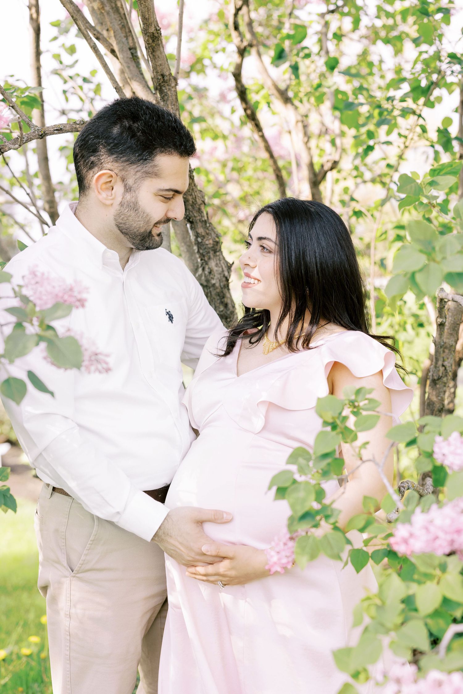 Loving expectant couple embracing among pink flowering trees, man tenderly holding pregnant womans belly.