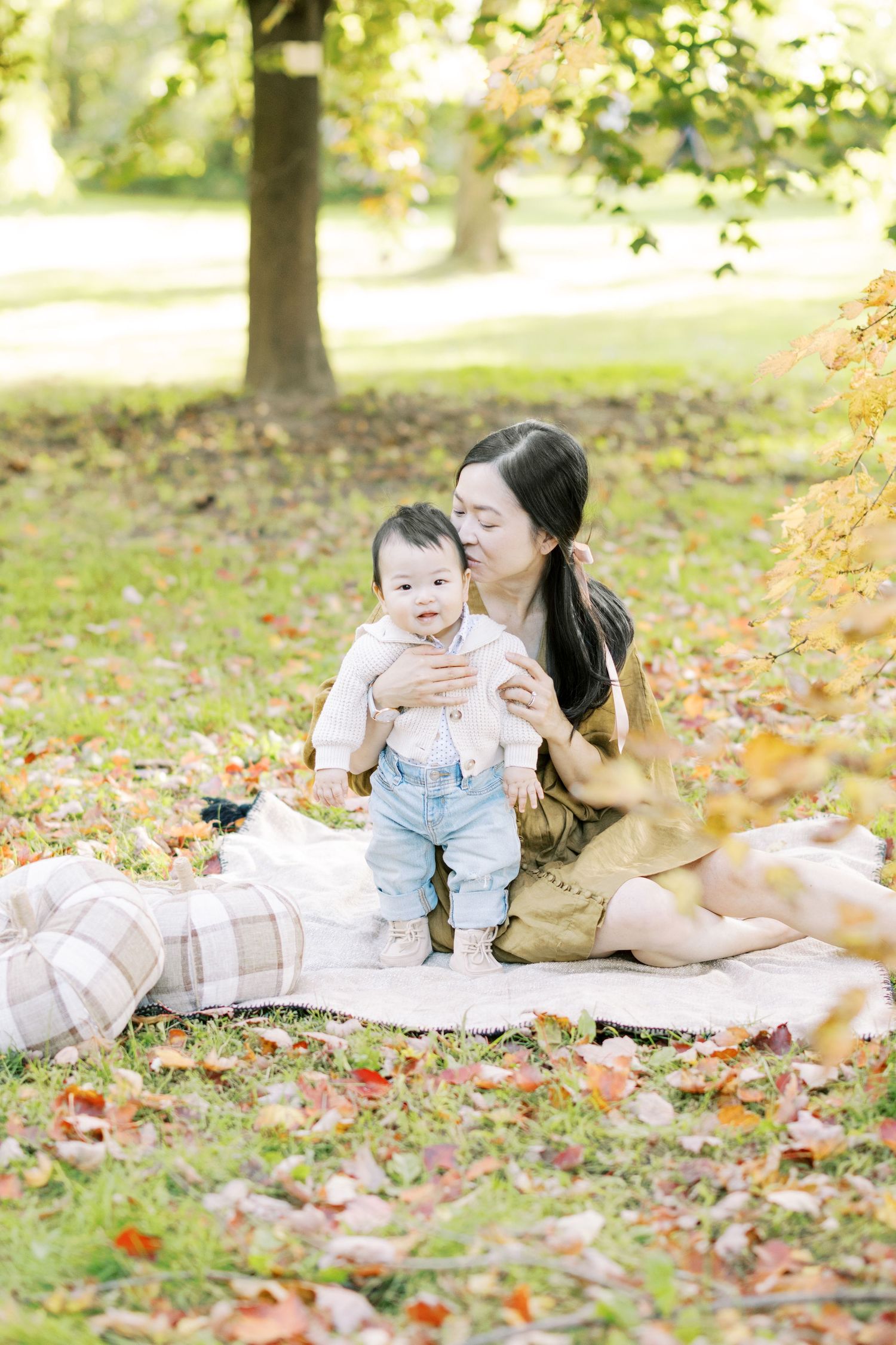 A mother kisses her baby while sitting on a blanket covered in colorful fall leaves.