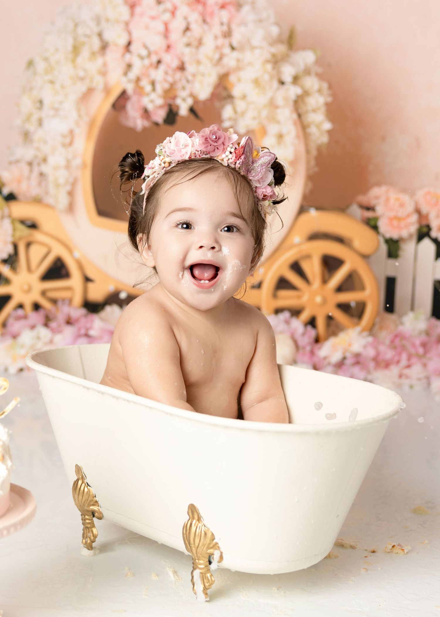 Joyful baby girl with pink flower crown splashes in white gold clawfoot tub at princess birthday photo shoot.