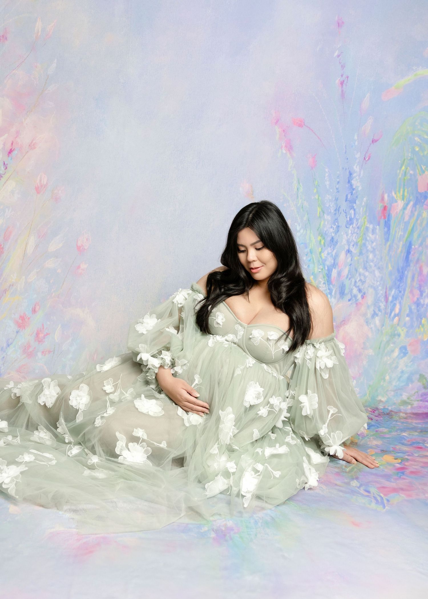 Pregnant woman in sage green floral tulle gown poses gracefully against a soft pastel painted backdrop.