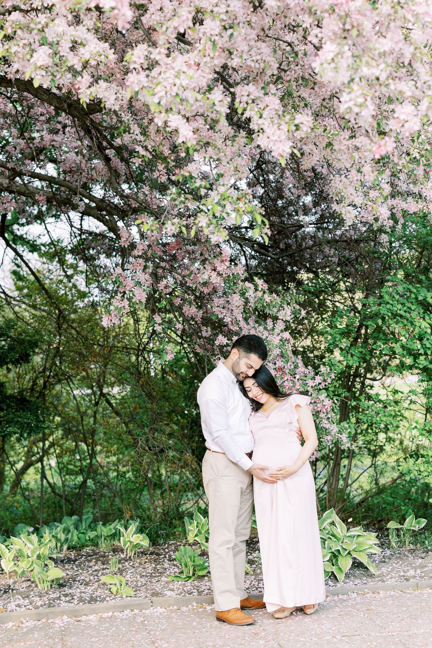 Pregnant couple embracing tenderly under pink blossoming tree in lush garden during spring maternity photo session.