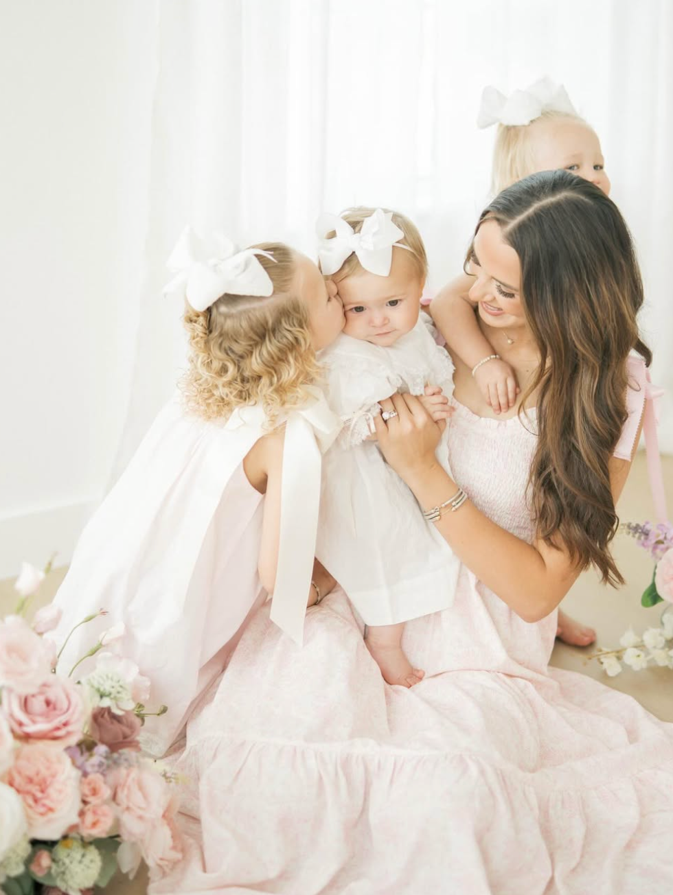 A mother in a pink dress smiles as her young daughters in white dresses and bows kiss a baby girl.