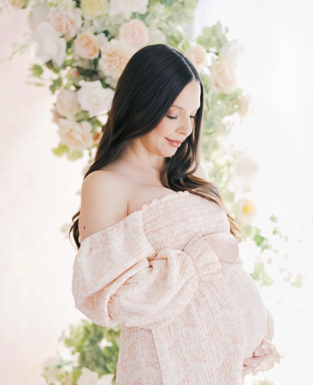 Pregnant woman in off-shoulder pink tweed dress poses gracefully before a floral backdrop of soft blush blooms.