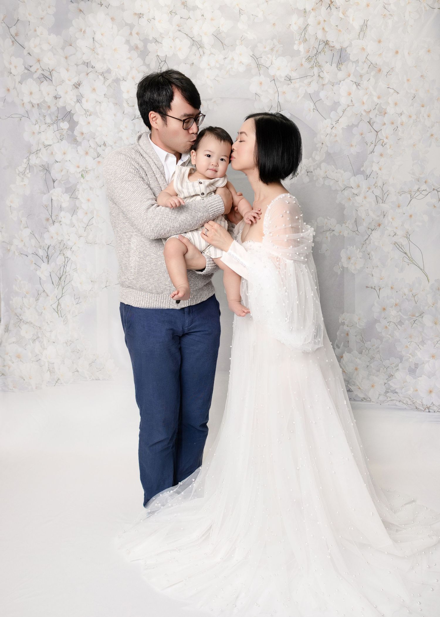 Asian couple in studio kissing baby, mother wearing elegant white gown on floral backdrop.