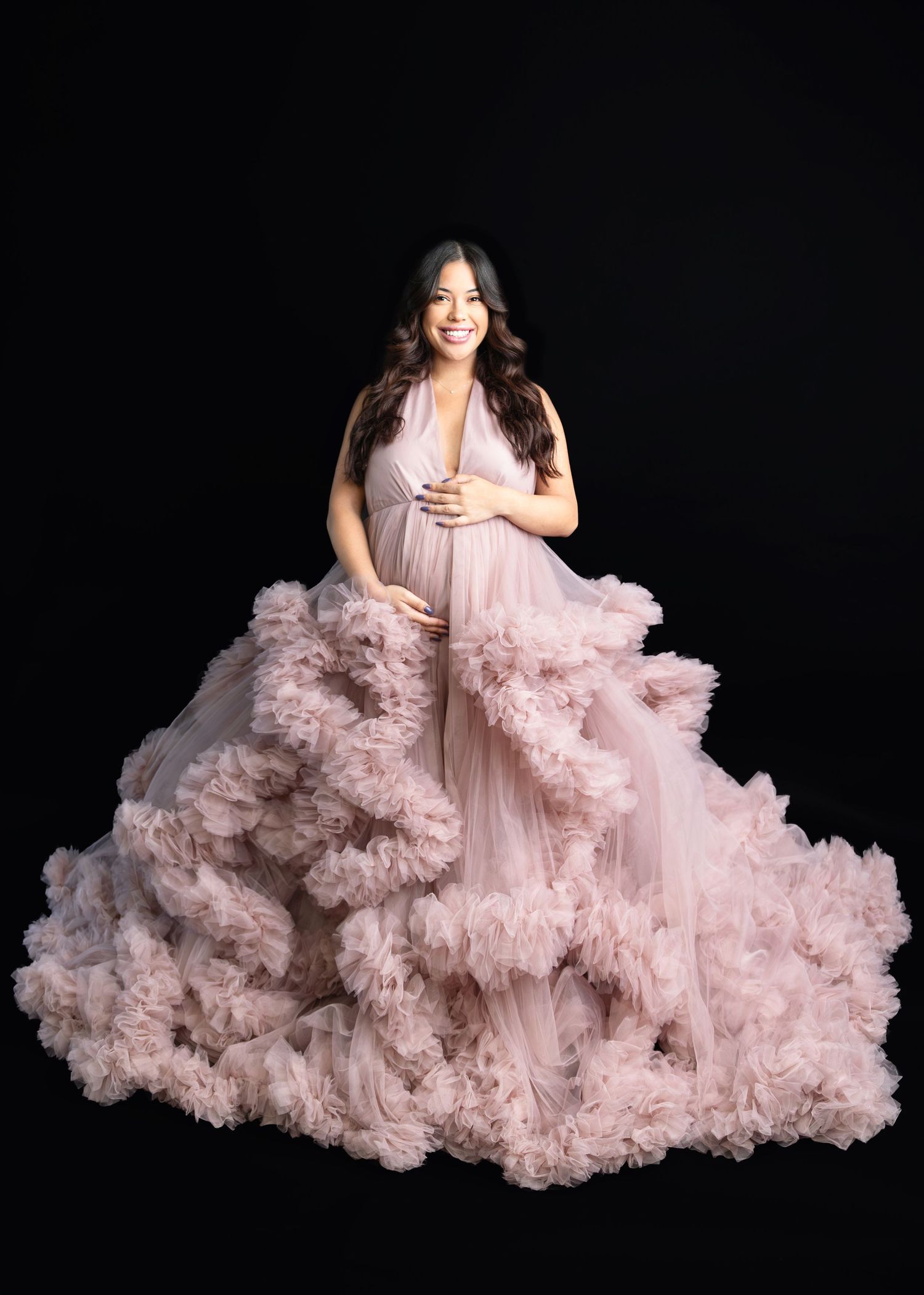Expecting mother in voluminous pink ruffle tulle gown cradling baby bump on black background.