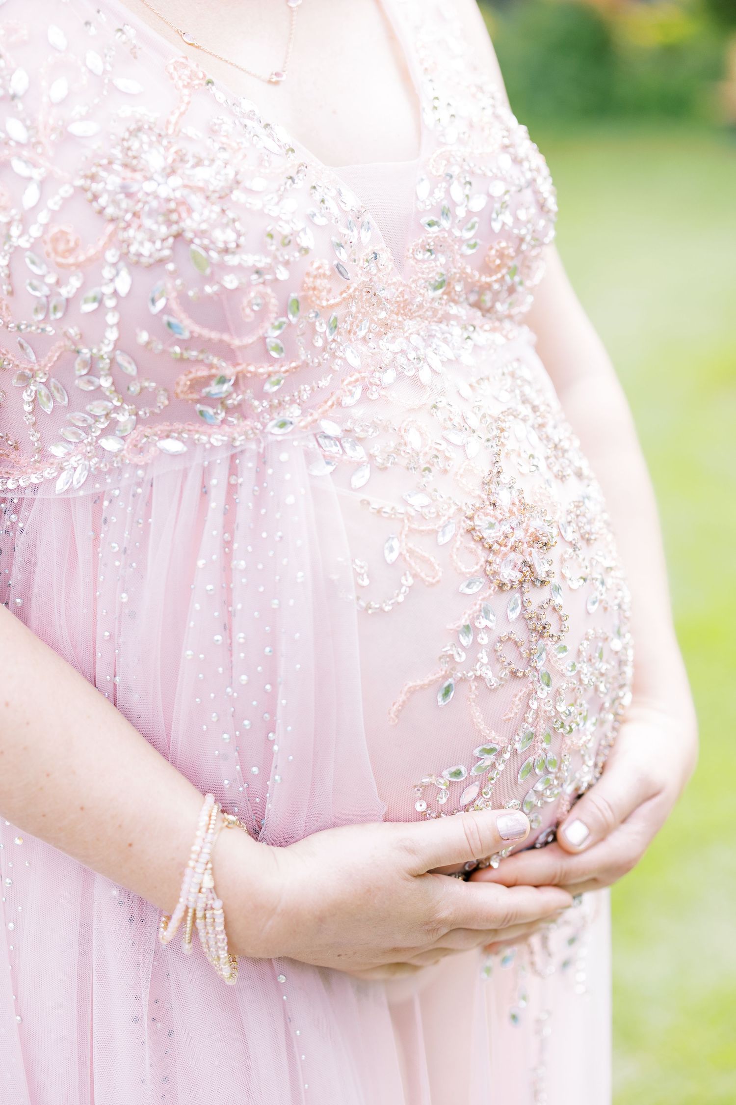 Close-up of pregnant belly in ornate pink beaded maternity gown with floral embroidery and pearl bracelet.