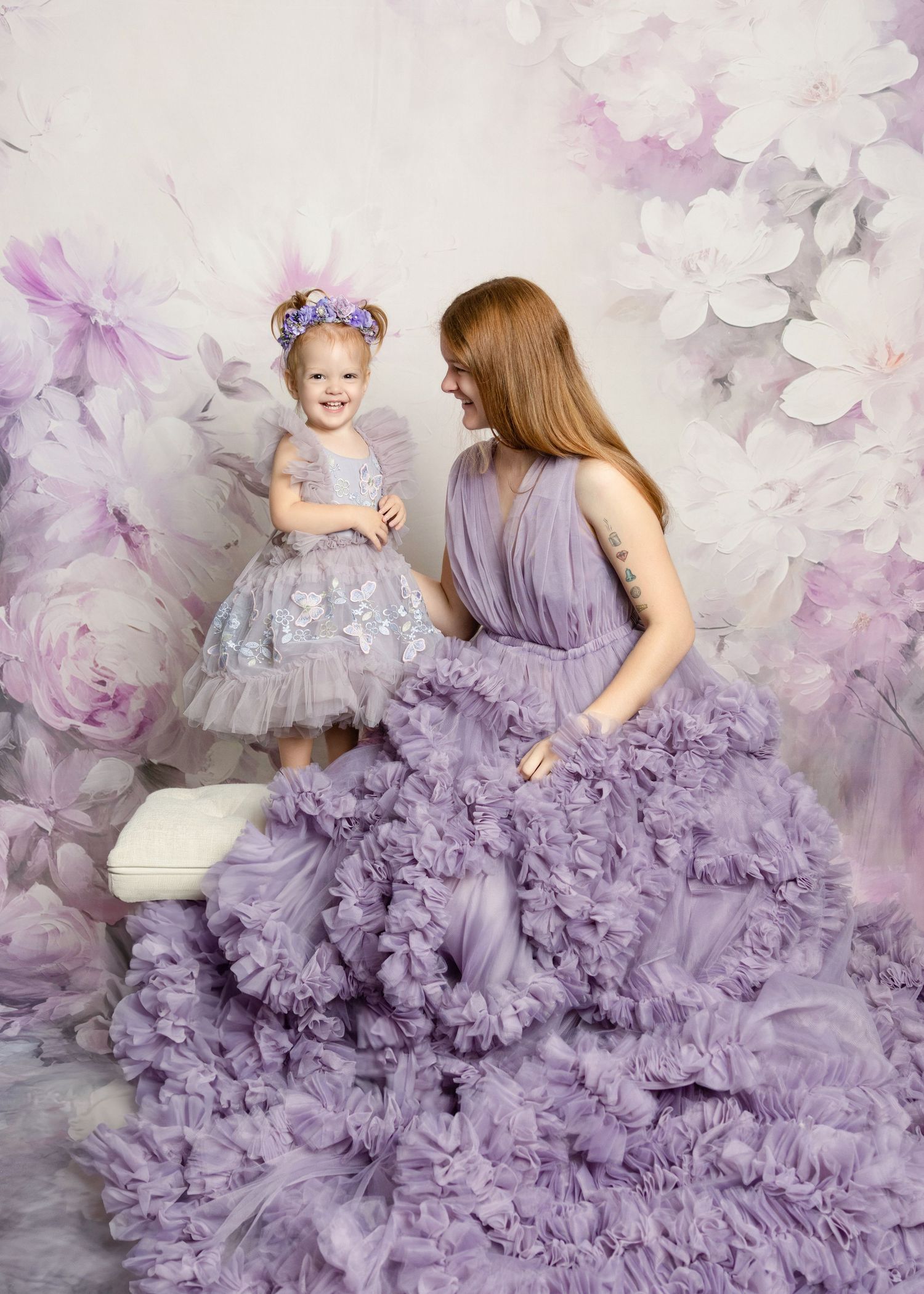 A mother and toddler daughter pose in matching lavender ruffle gowns against a floral backdrop.