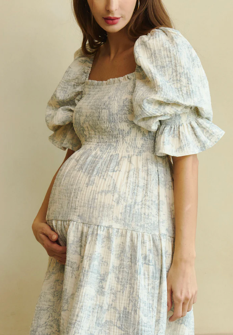 Pregnant woman wearing a floral smocked bodice midi dress with puff sleeves and tiered skirt in soft blue and cream.