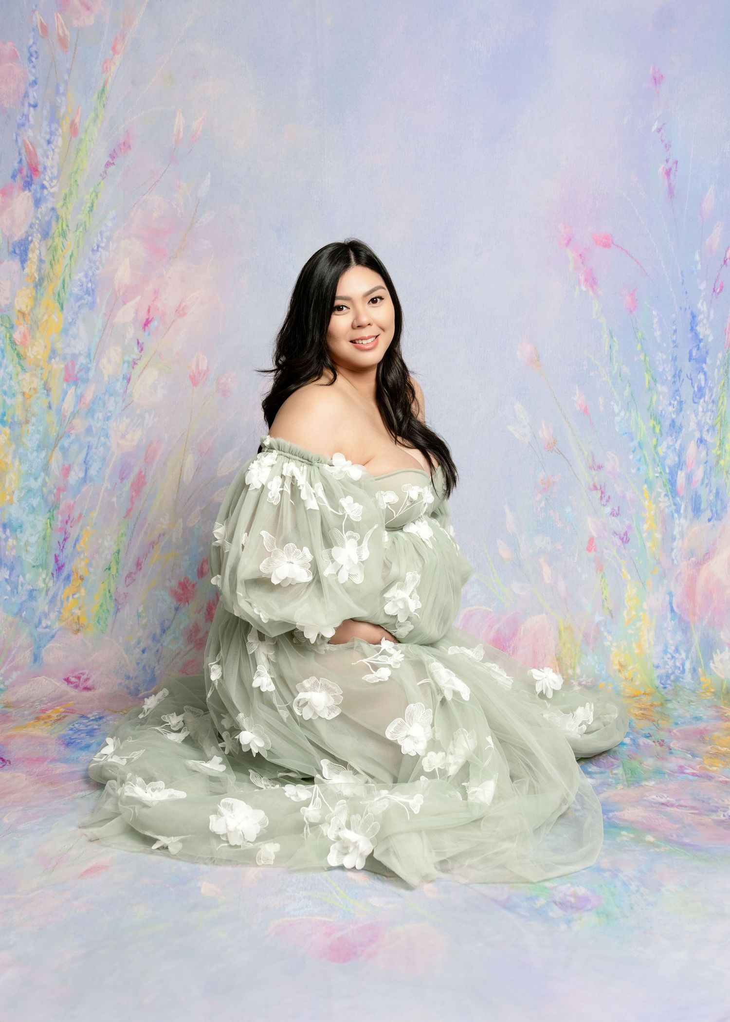 Woman in sage green floral tulle gown sits gracefully before a soft pastel painted backdrop.