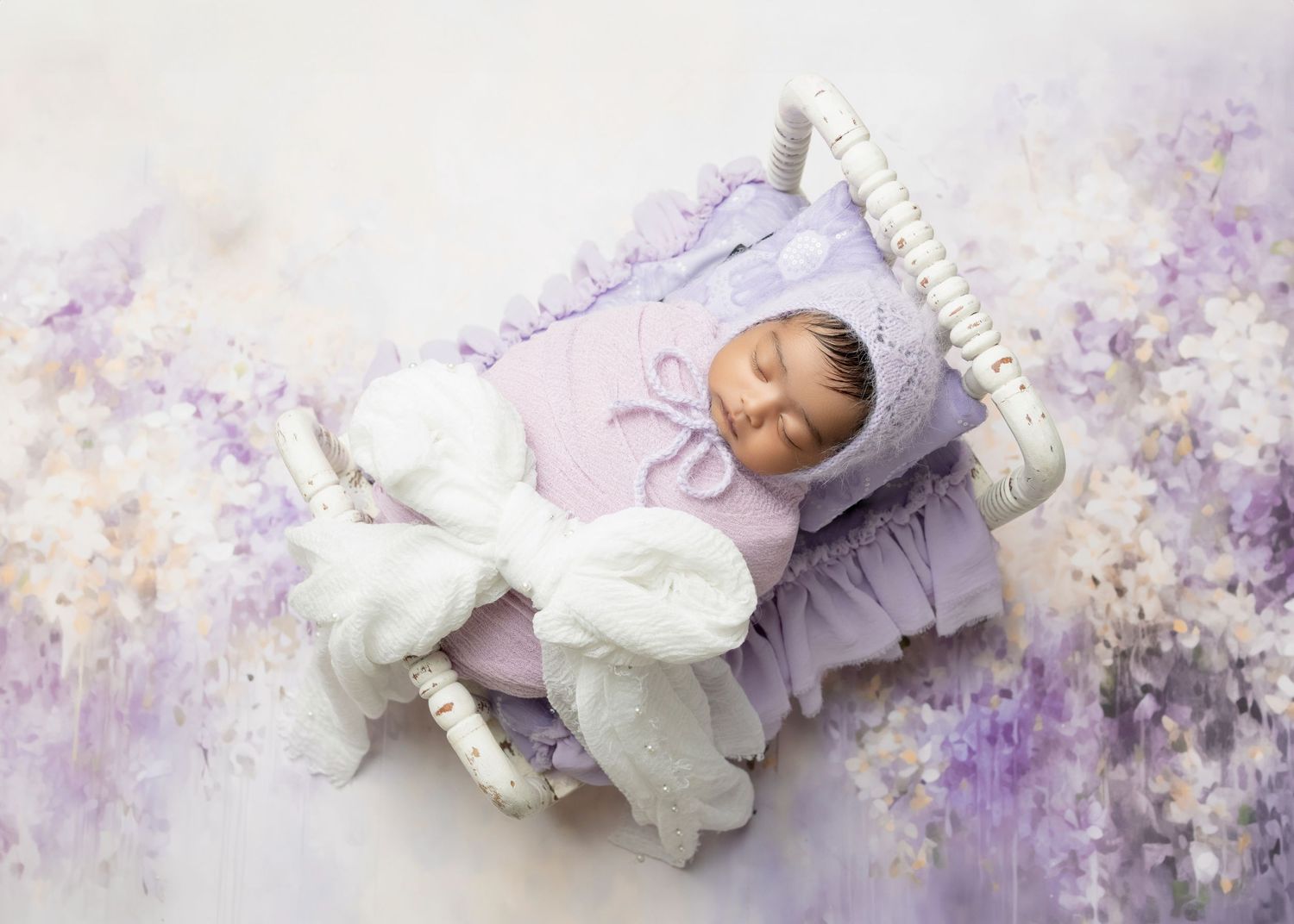 Newborn baby sleeping peacefully in a white mini bed with purple bedding and a white stuffed bunny on a floral background.
