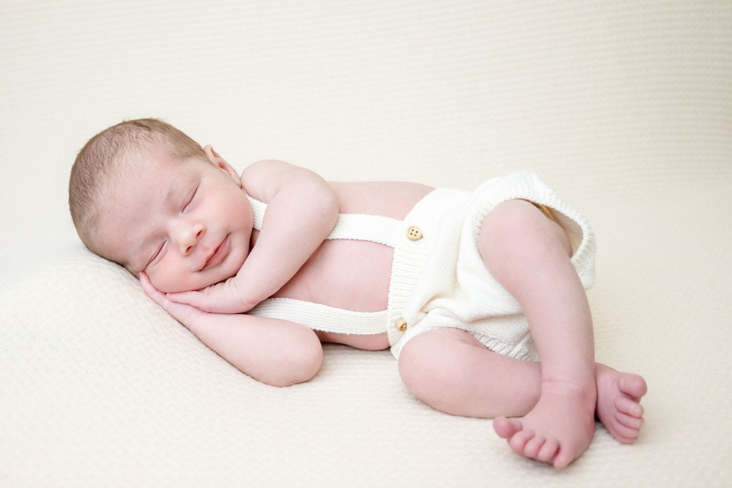 Smiling sleeping newborn baby in white suspender onesie lying on cream blanket in professional portrait photo.