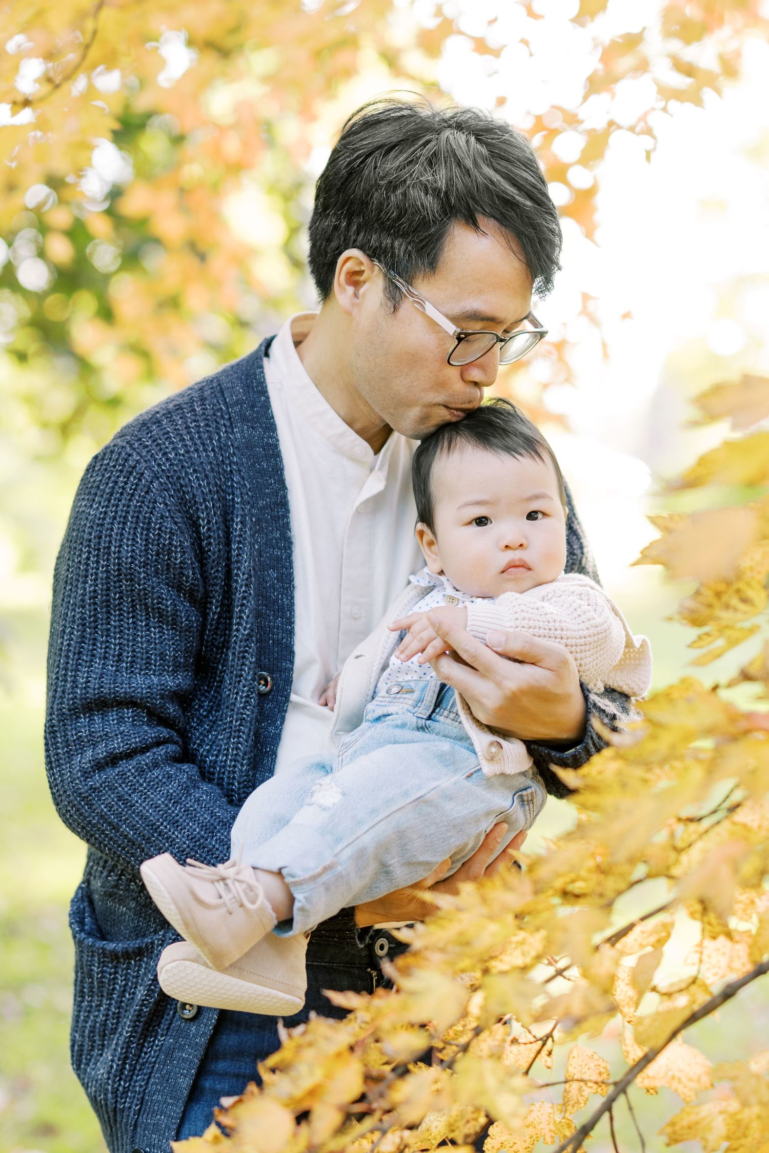 A father wearing glasses kisses his baby girl amid golden autumn leaves in a park.