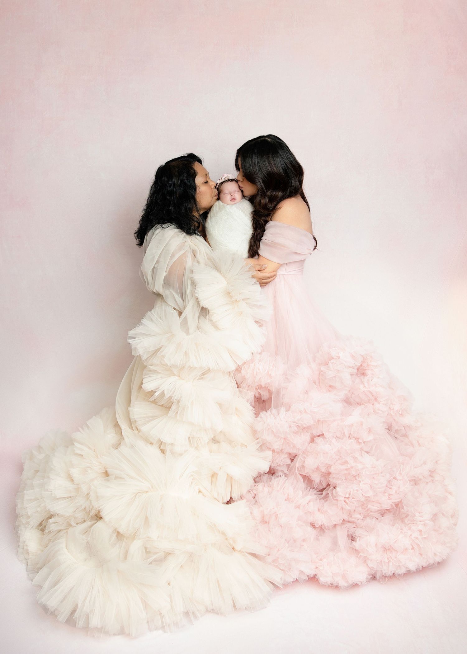 Two women in white and pink tulle gowns kissing a sleeping newborn baby between them.