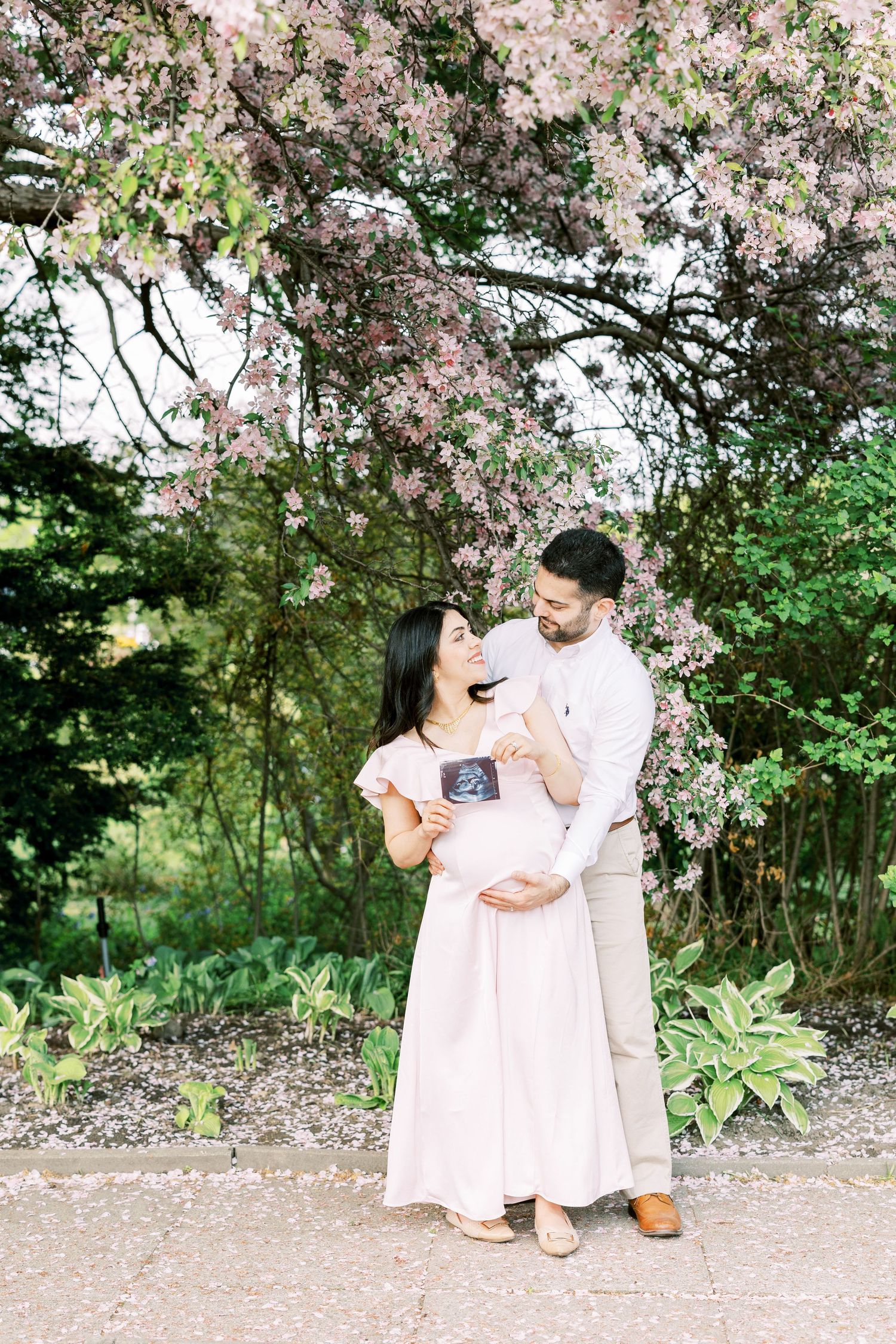 Happy expecting couple sharing tender moment under blooming pink tree holding baby ultrasound image.