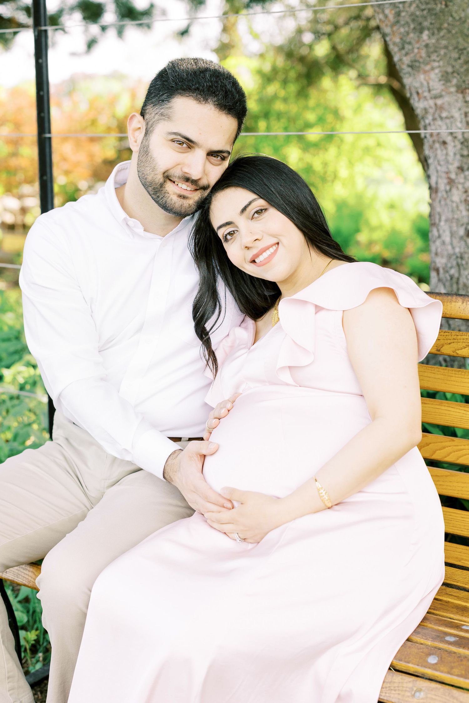 Smiling pregnant couple sitting on wooden bench in park, hands gently cradling baby bump.