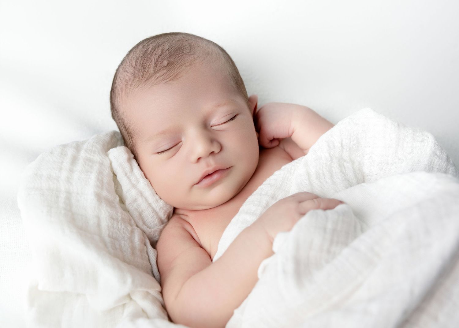 Peaceful newborn baby sleeping wrapped in soft white muslin cloth on a white background.