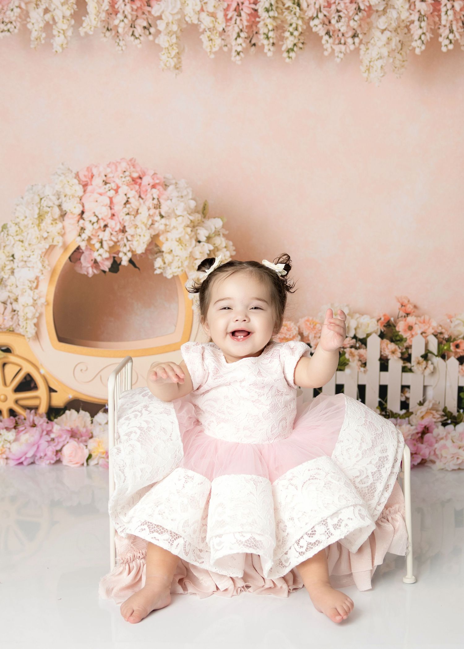 Smiling baby girl in white lace pink dress sits in miniature bed with floral backdrop and carriage prop.