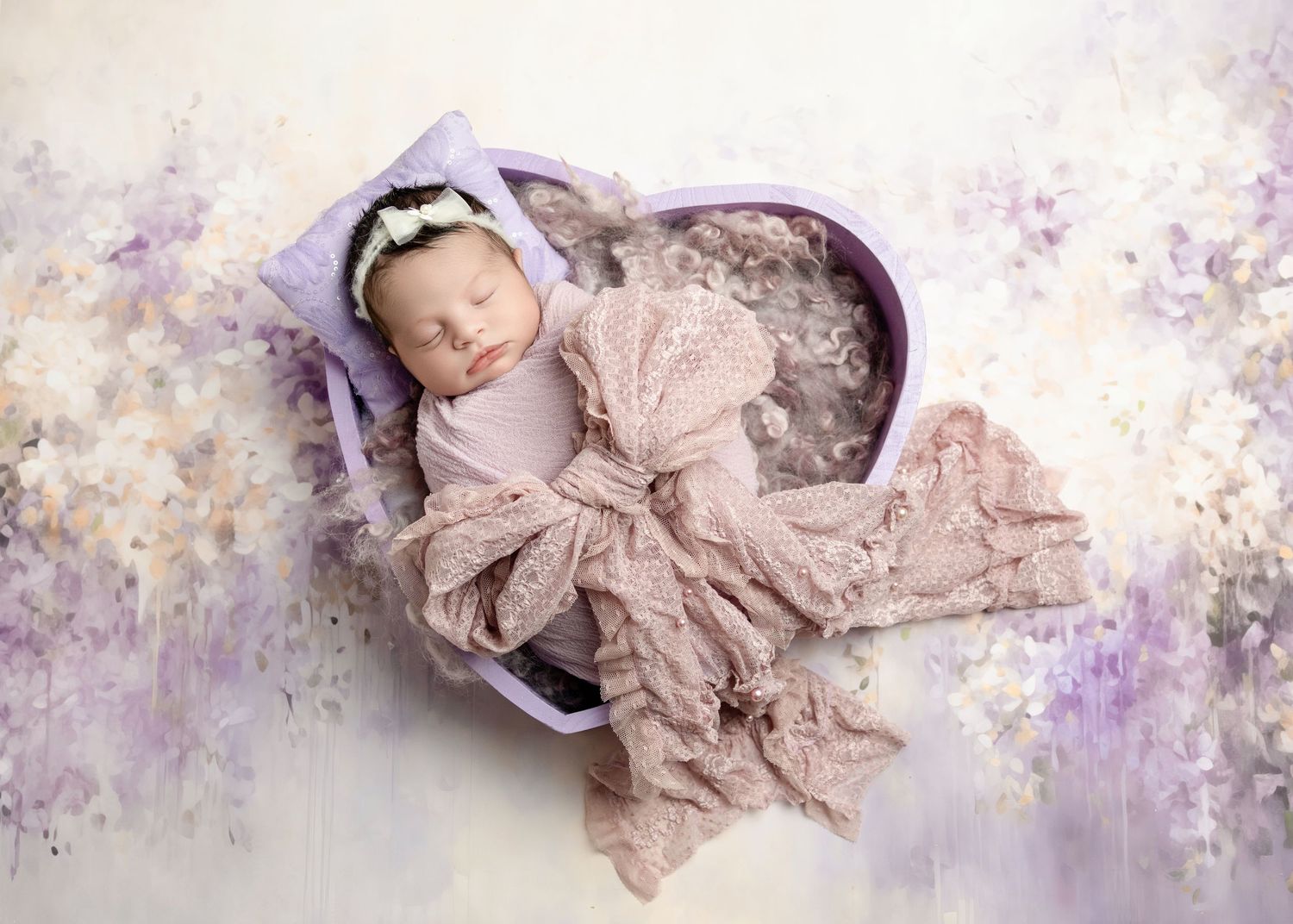 Newborn baby girl wrapped in lavender lace sleeping in a heart-shaped box with floral backdrop.