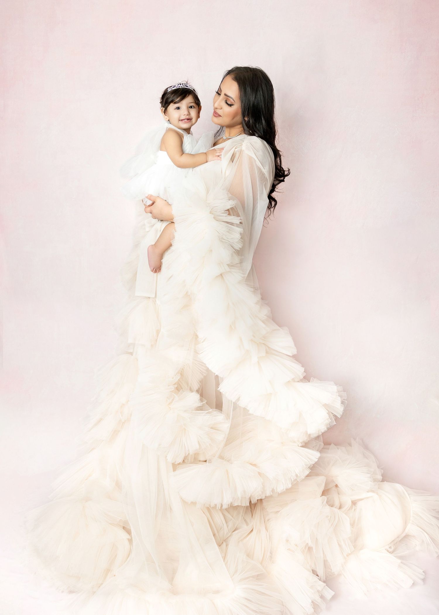 Elegant mother in white ruffled gown holding young daughter against soft pink studio background.