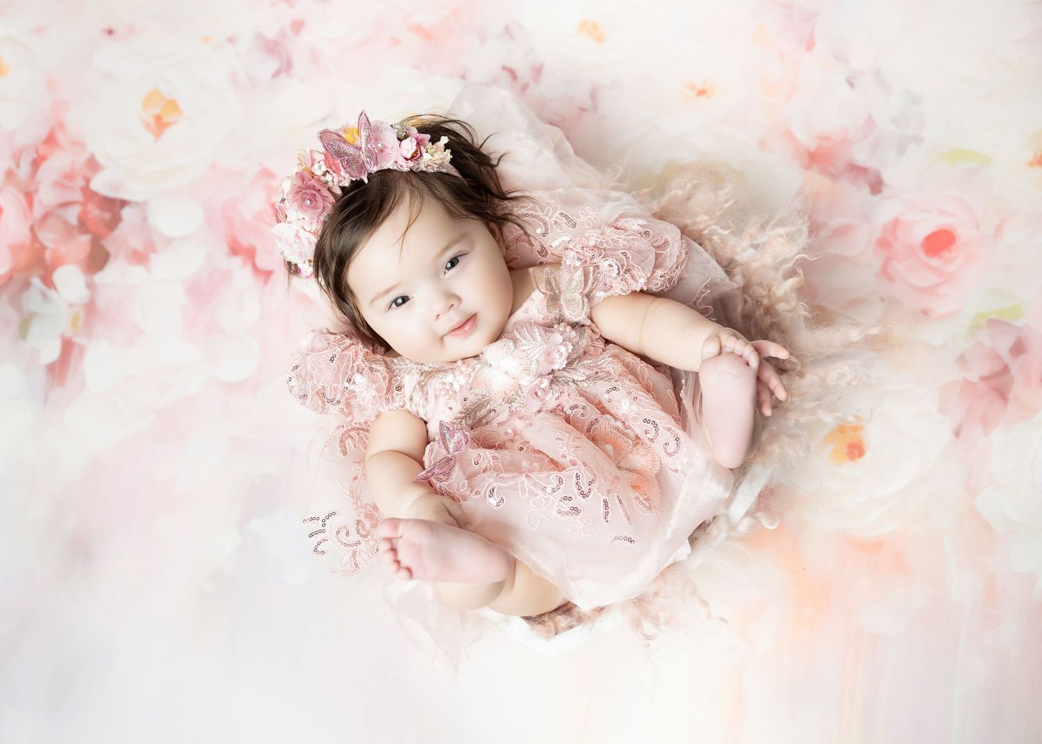 Baby girl in pink lace romper lying in a floral bowl arrangement from above.