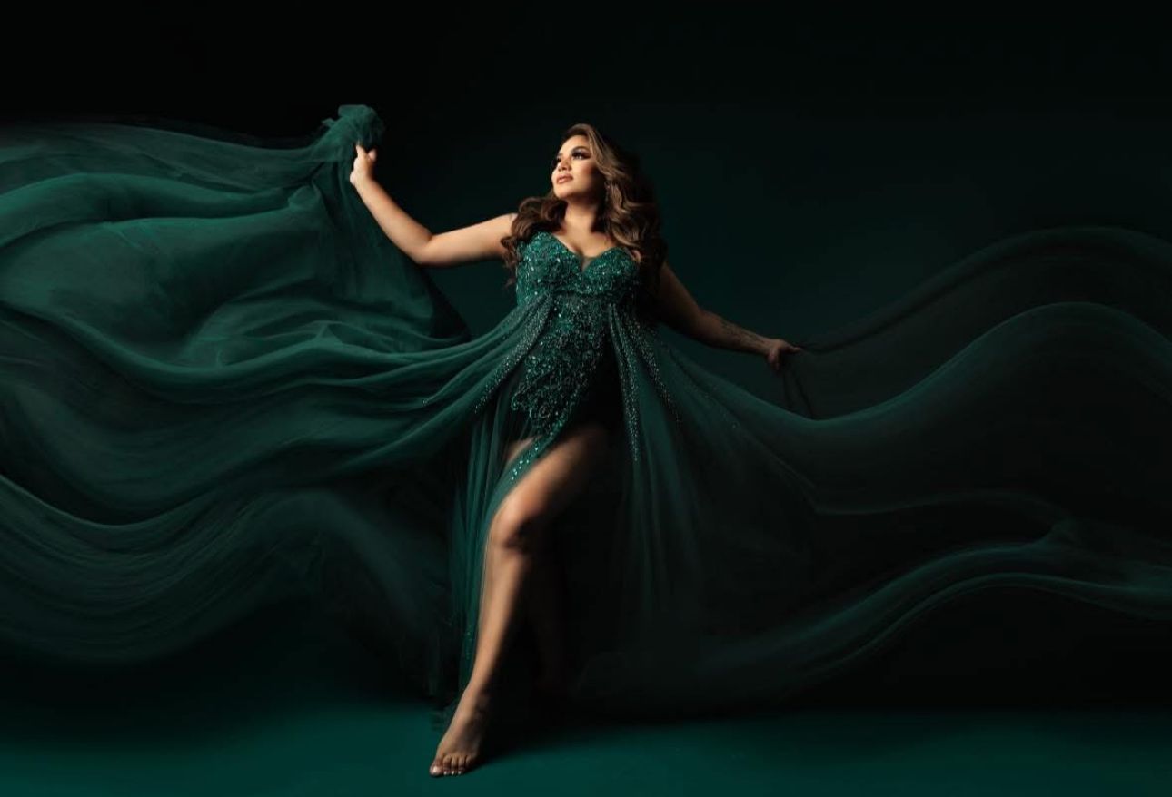 Woman in flowing emerald green beaded gown with dramatic fabric wings poses gracefully against dark background.