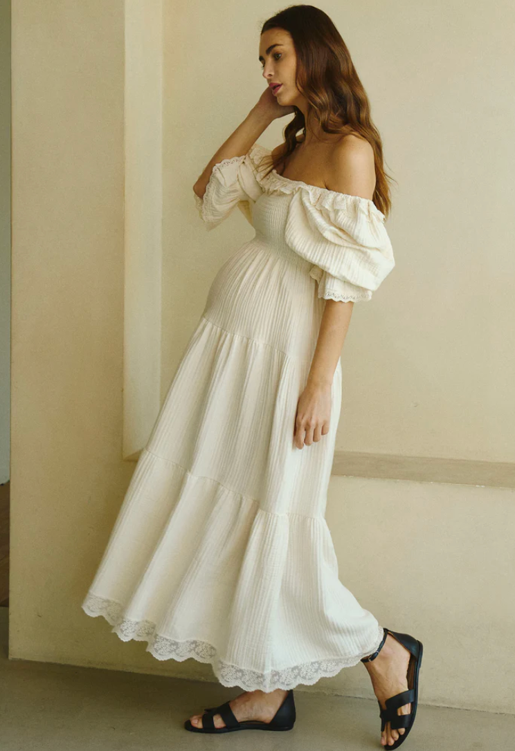 Woman wearing a white off-shoulder maxi dress with puff sleeves, lace hem, and black sandals.