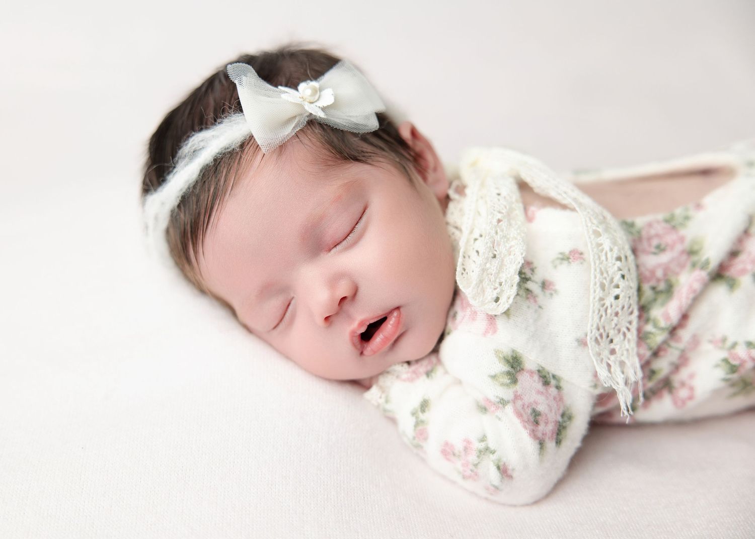 A close-up of a sleeping newborn baby girl in a floral outfit with a delicate white bow headband.