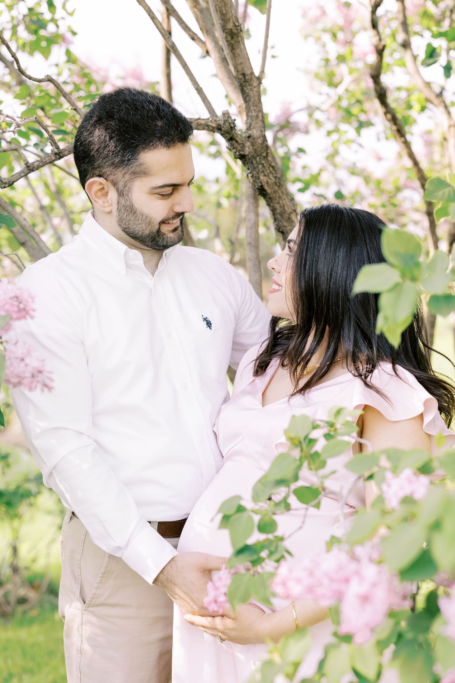 A couple shares a tender moment among pink blossoms, the woman in a pink dress cradling her baby bump.
