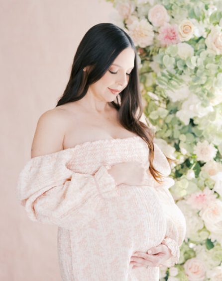 Pregnant woman in blush pink off-shoulder gown cradling baby bump before floral wall backdrop.
