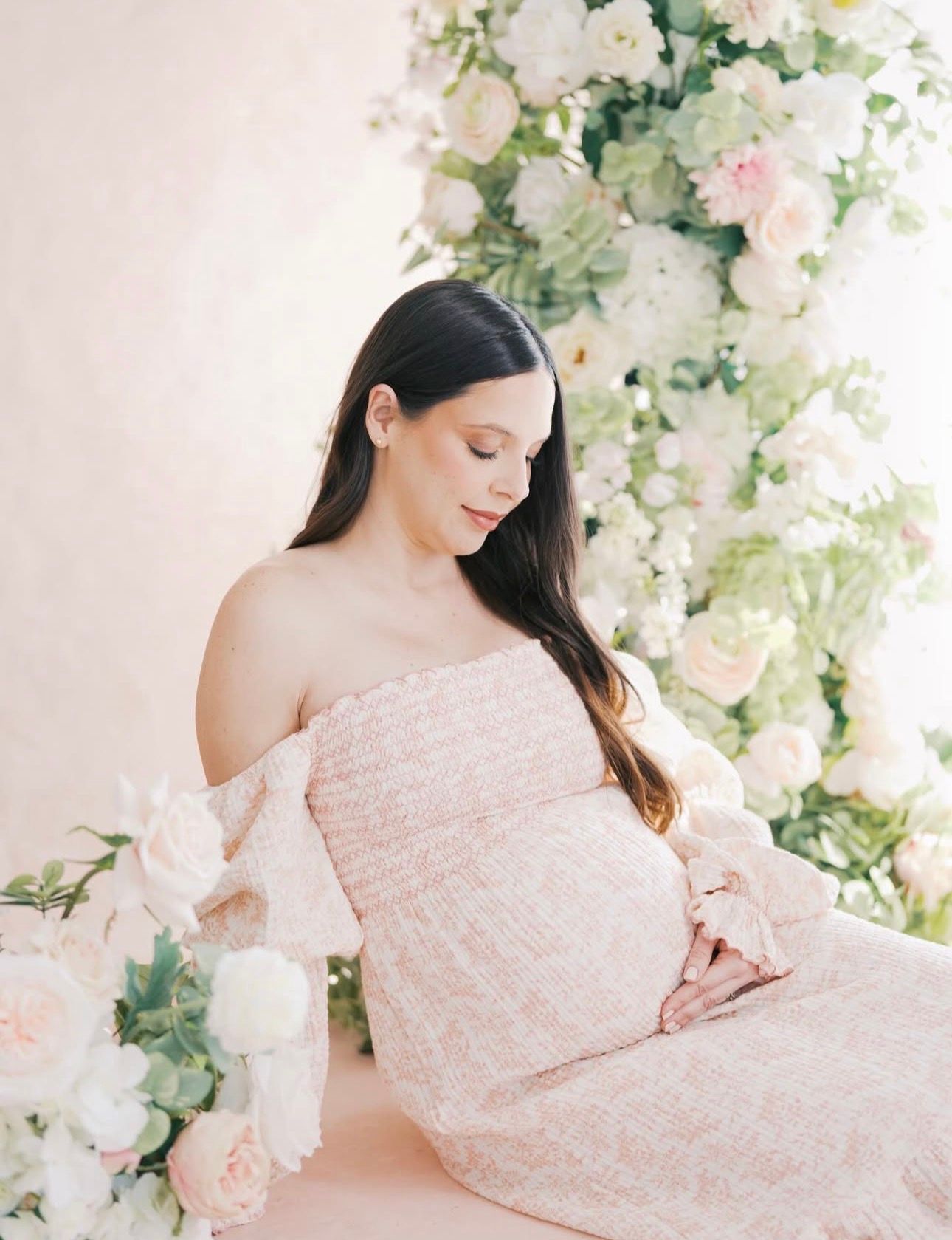 Pregnant woman in pink off-shoulder dress poses gracefully among lush white and pink floral arrangements in a soft studio setting.