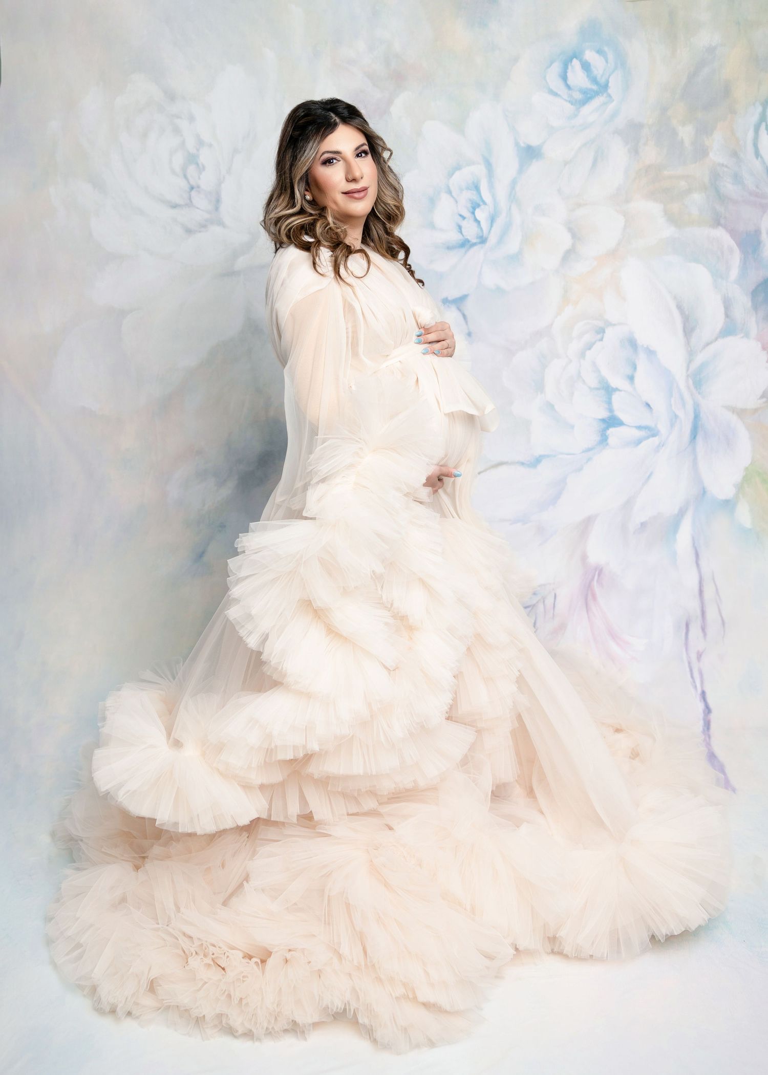 Pregnant woman in elegant blush tulle gown poses gracefully against a soft floral painted backdrop.