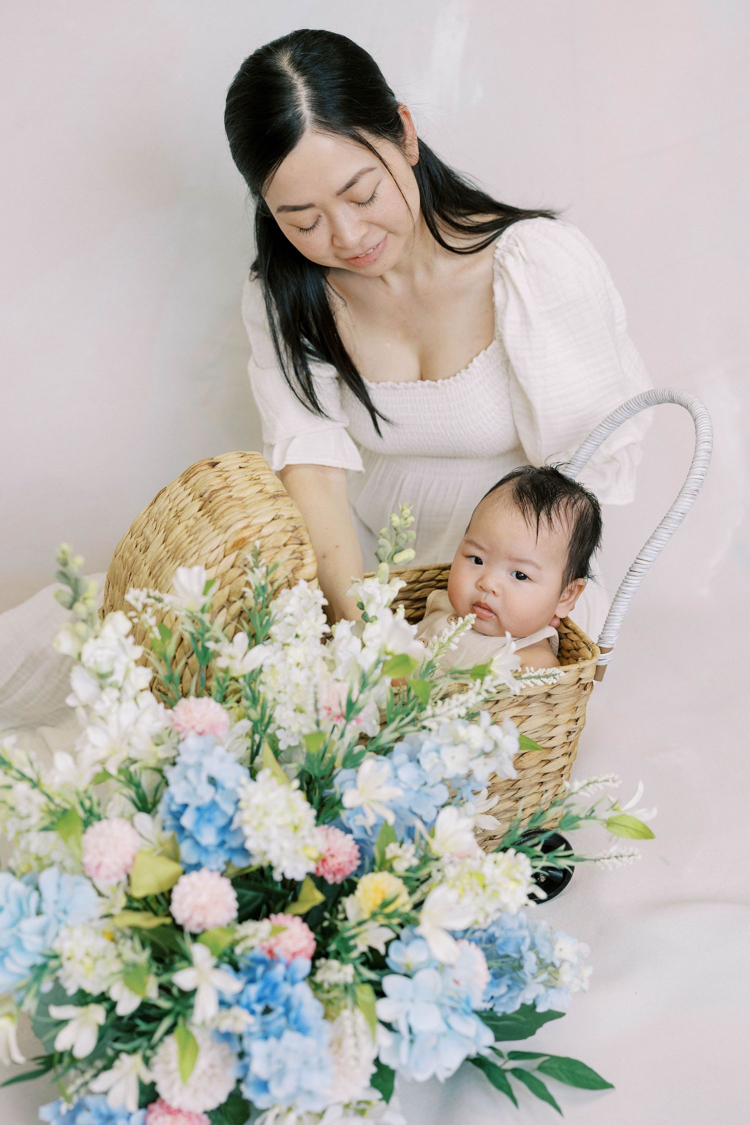 Mother in white dress smiling at baby sitting in wicker basket surrounded by blue and white floral arrangement.