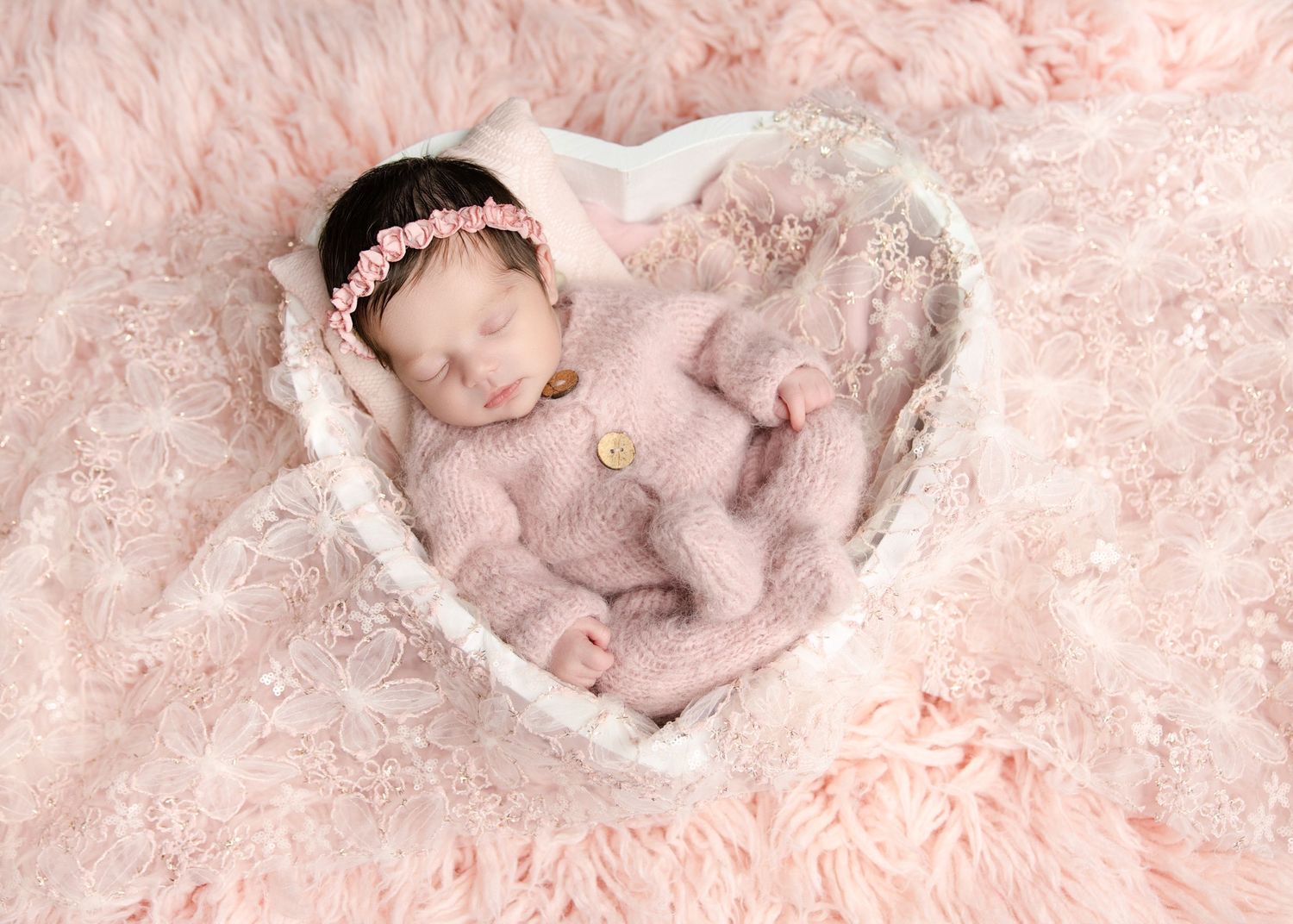 A sleeping newborn baby girl in a pink fuzzy romper rests in a heart-shaped basket on pink fur.