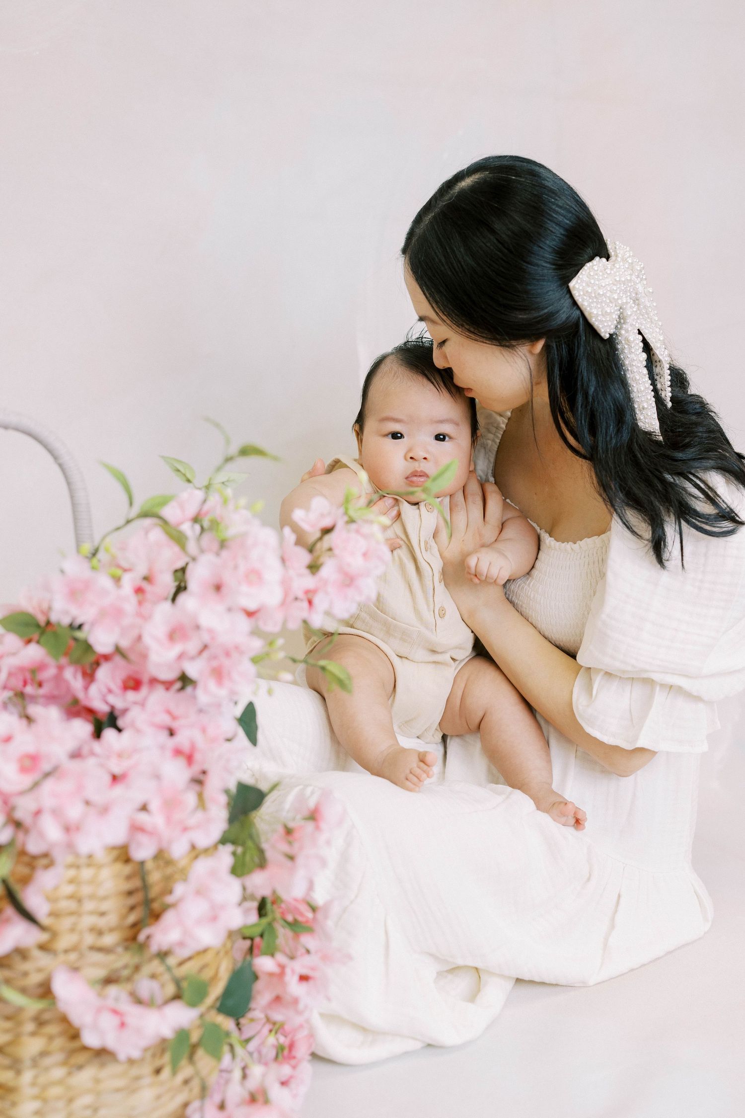 Mother in white dress kissing baby on cheek while seated beside basket of pink cherry blossom flowers.