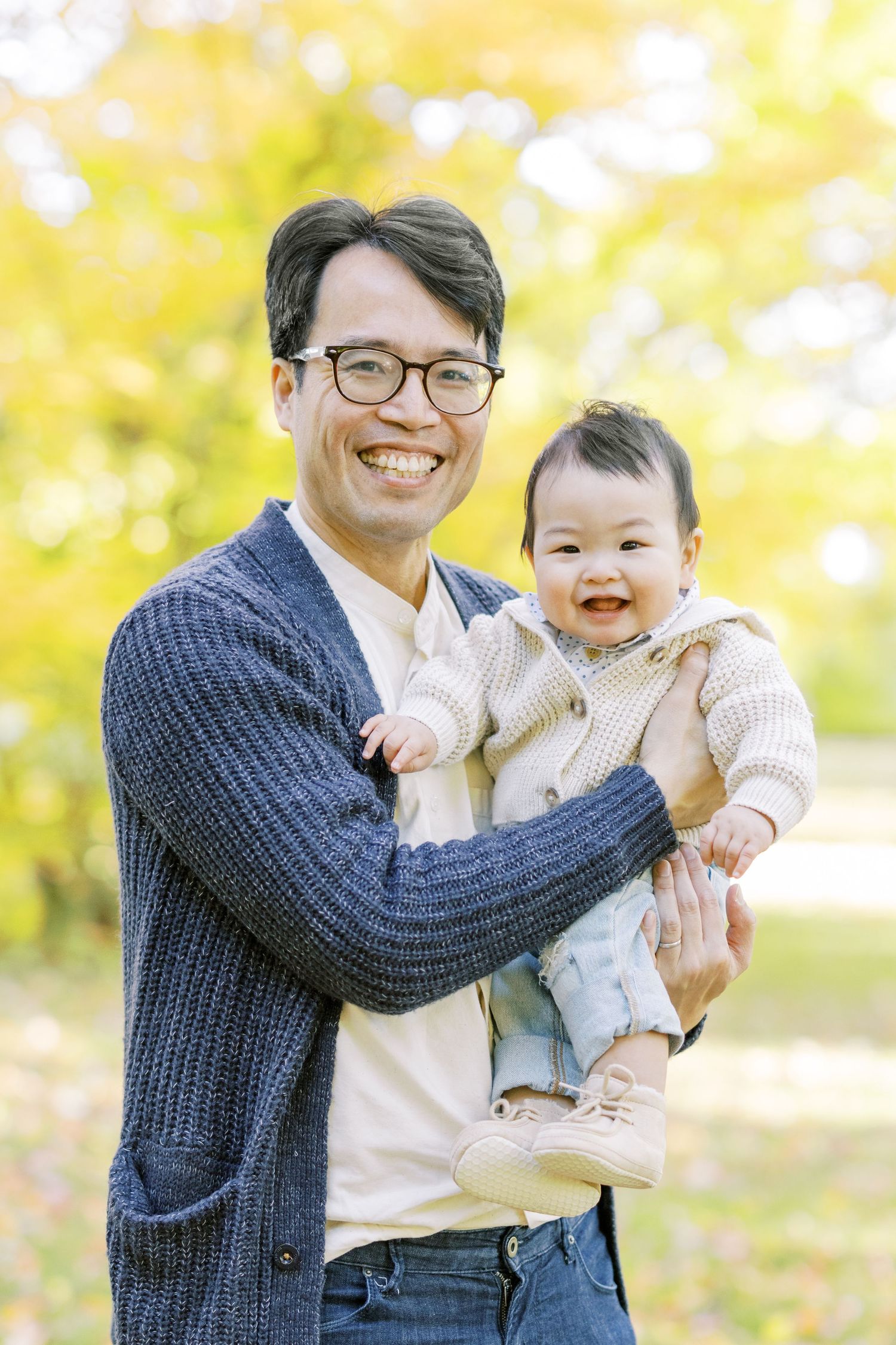 Smiling Asian father holding happy baby in autumn park with yellow foliage background.